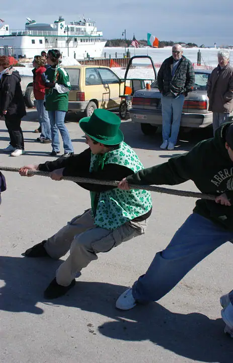 People pull a rope in a tug-of-war beside parked cars and a large ferry.