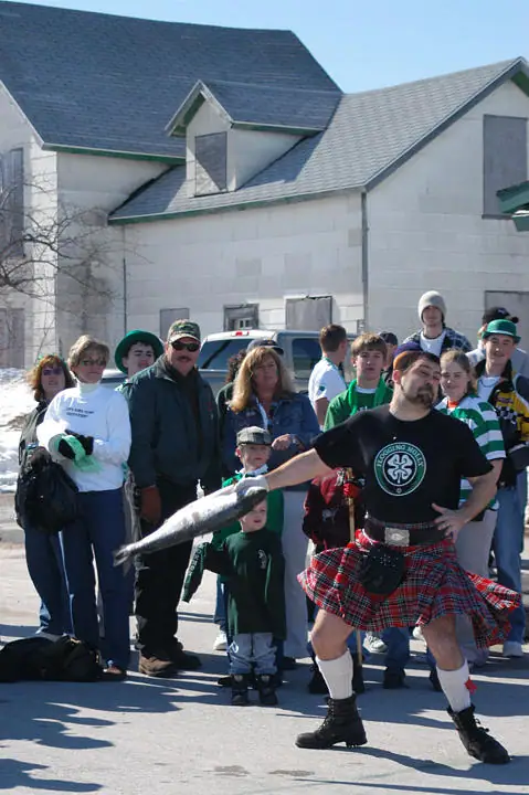 A man in a tartan kilt holds a large fish while a crowd gathers outside a building.