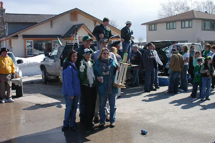 People in green clothing gather outdoors beside snowbanks and vehicles.