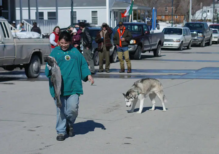 A child carries a large fish across a paved street as a leashed dog follows nearby.