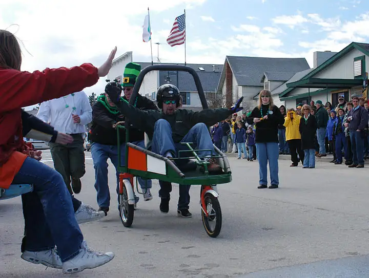 People pull a green-and-orange pedal cart along a street as spectators watch beneath flags.