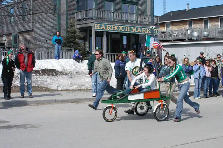 People push a decorated cart along a street past a storefront, with flags, spectators, and patches of snow visible.