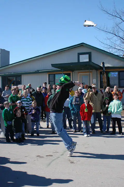 A person in a green hat jumps beneath a fish-shaped object as spectators watch outside a building.