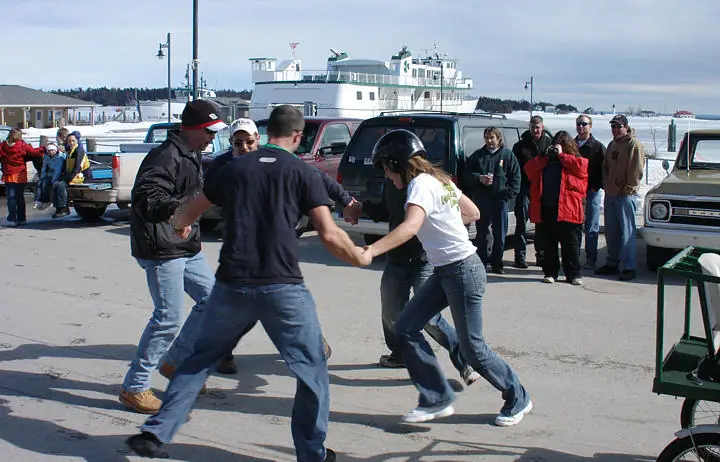 People take part in an outdoor game beside a ferry, with spectators, vehicles, and snow-covered water behind them.