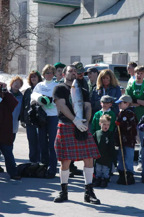 A kilted man holds a large fish while adults and children gather outside a building.