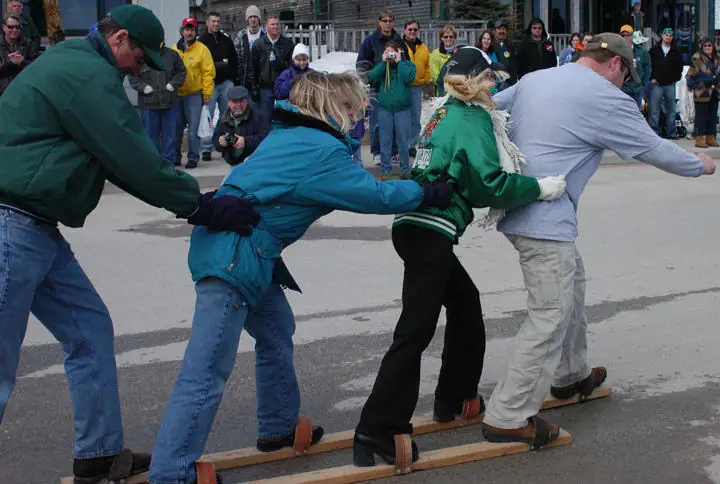 Four adults balance on parallel wooden planks while spectators watch and photograph an outdoor game.