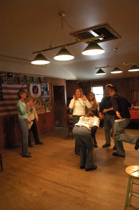 Several people dance in a wood-paneled room beneath pendant lights, beside a large American flag.