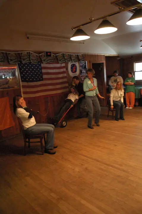 People gather inside a wood-paneled room, with one adult pushing a child in a stroller beneath a large American flag.