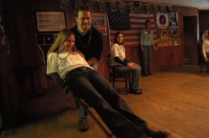 People gather in a wood-paneled room as a seated woman leans back in a chair during a playful moment.