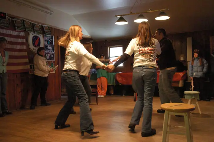 People dance in a wood-paneled room decorated with an American flag, beer signs, and colorful tables.