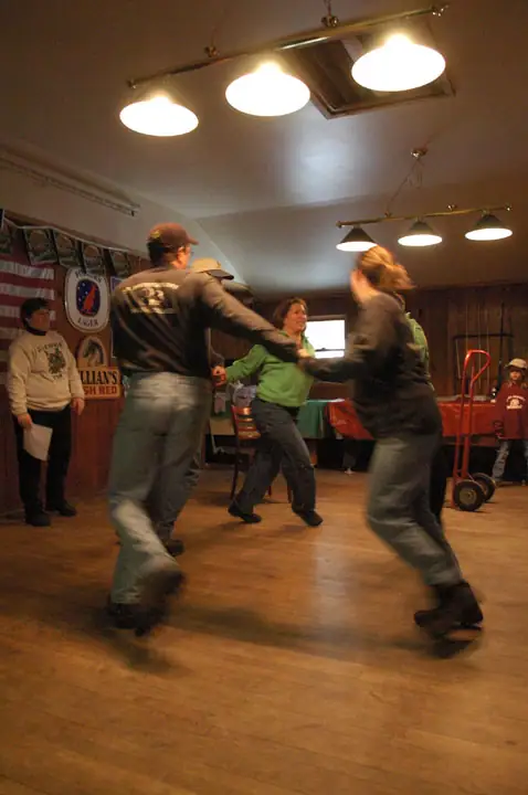 Adults dance on a wooden floor while others watch in a brightly lit room with beer posters and a hand truck.