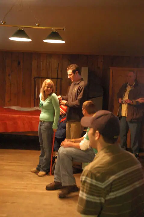 Several people gather in a wood-paneled room beneath hanging lights, with some seated and others standing near a red-covered table.