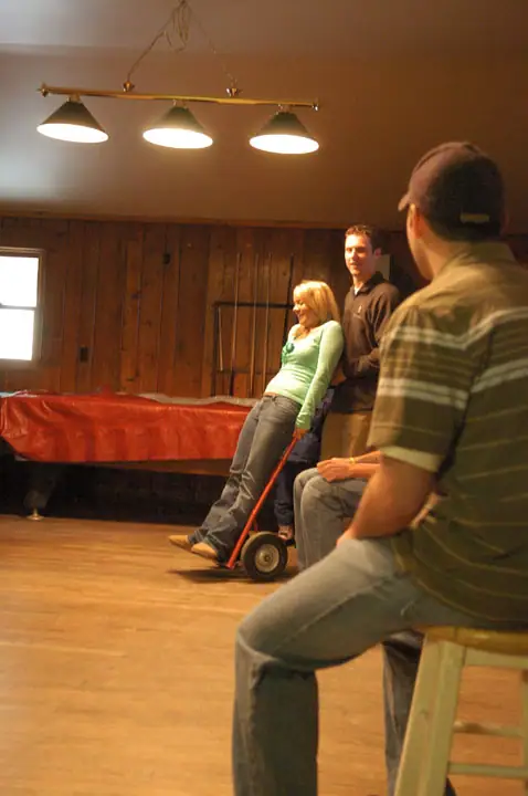 Three people gather in a wood-paneled room beside a red-covered table and a hand truck.