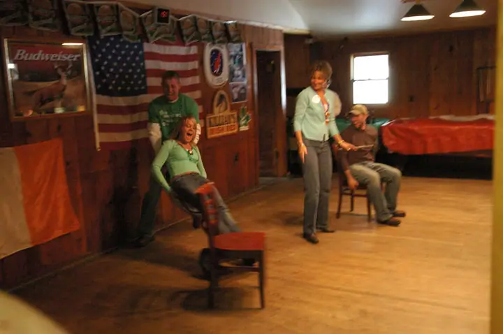 Four adults gather in a wood-paneled room with flags, posters, chairs, and a Budweiser advertisement.