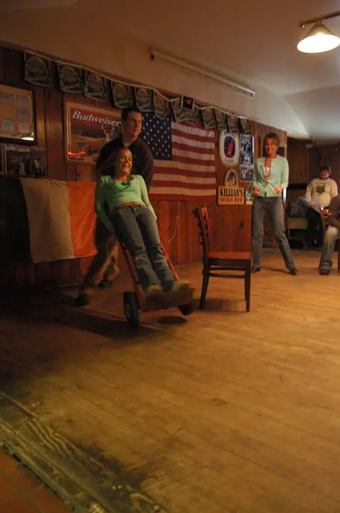 People gather indoors as a woman rides in a red wheeled cart beneath Irish and American flags.