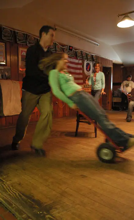 A man pushes a seated woman on a small red wheeled cart across a wood-paneled room.