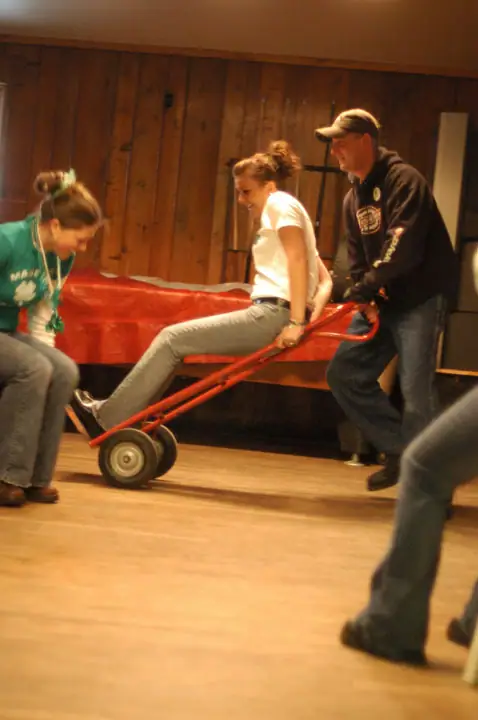 Three people playfully move a seated woman on a red hand truck inside a wood-paneled room.