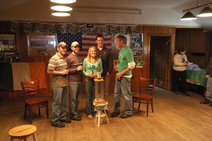 Five adults pose around a small stand in a wood-paneled room decorated with flags, banners, and green holiday decorations.