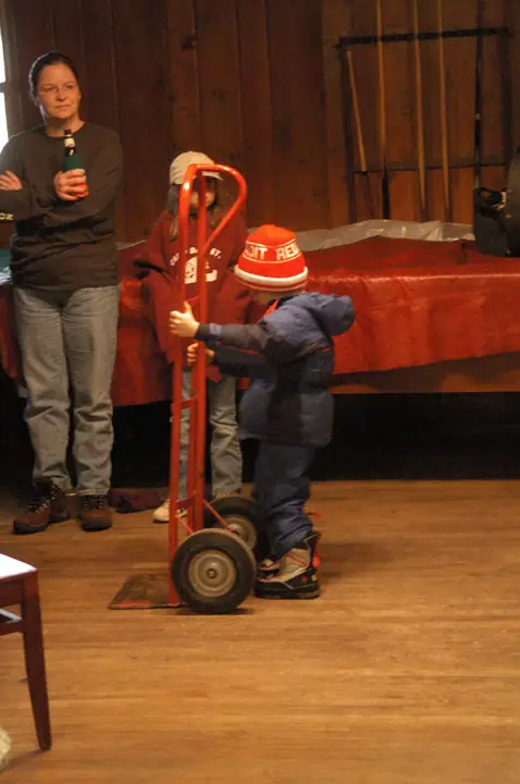 An adult and two children stand indoors beside a red hand truck and a table covered with a red cloth.