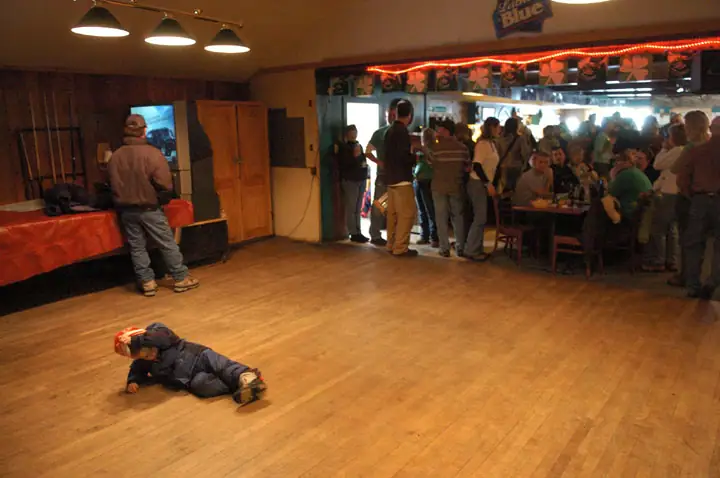 A child lies on a wooden floor while people gather near a decorated bar and tables.