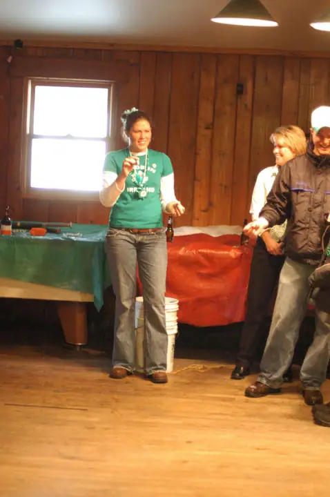 A woman gestures while three people stand in a wood-paneled room beside green- and red-covered tables.