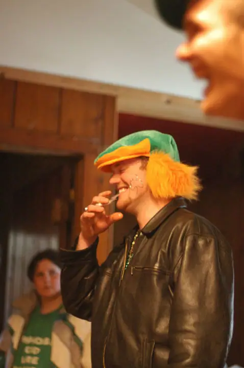 A person in a green-and-orange novelty hat and black leather jacket holds a cigar near their mouth indoors.