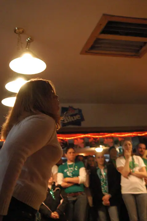 A woman stands in profile before a group of people gathered indoors under bright lights and red string lighting.
