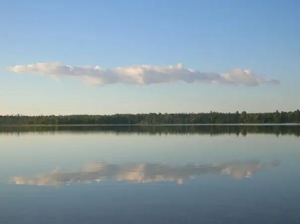 Still water reflects a tree-lined shore and a long band of clouds beneath a clear blue sky.