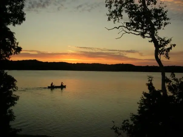 A canoe with two silhouetted occupants crosses calm water at sunset, framed by dark trees.