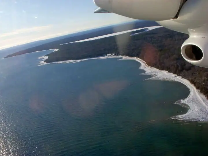 Aerial view of a wooded shoreline with snow or ice, seen beside an aircraft engine.