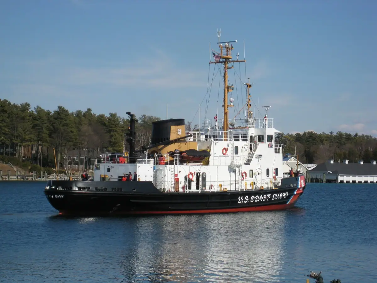 A Coast Guard cutter travels past docks and a wooded shoreline.
