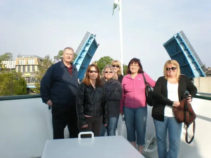 Six adults pose on a passenger vessel deck with a raised drawbridge and waterfront buildings behind them.