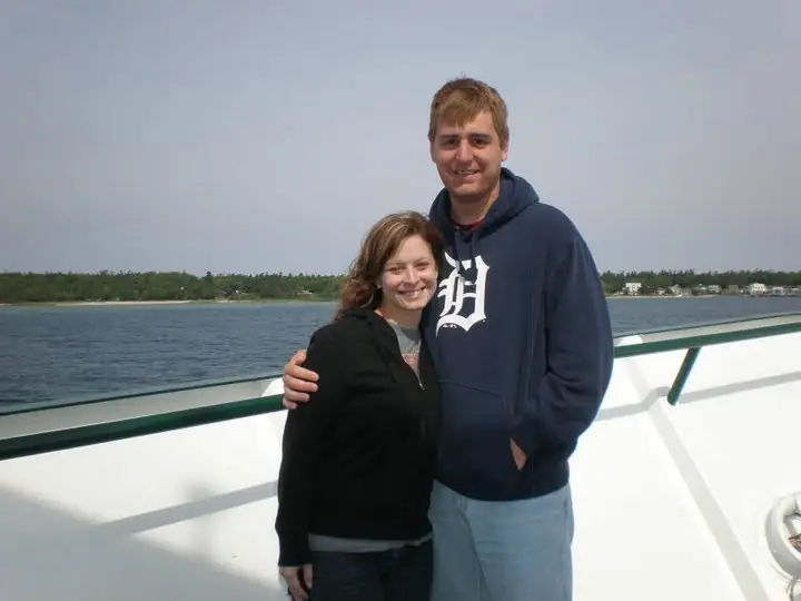 A young couple stands together on the open deck of a passenger vessel with a wooded shoreline behind them.