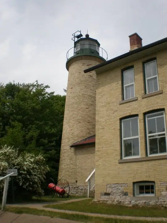 A brick lighthouse tower and adjoining brick building stand beside trees beneath an overcast sky.