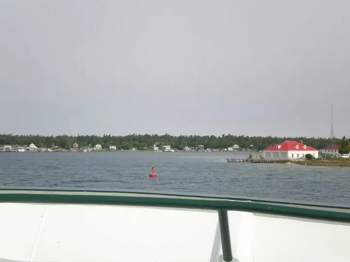 View across open water from a boat deck toward a wooded shoreline, waterfront buildings, a pier, and a red buoy.