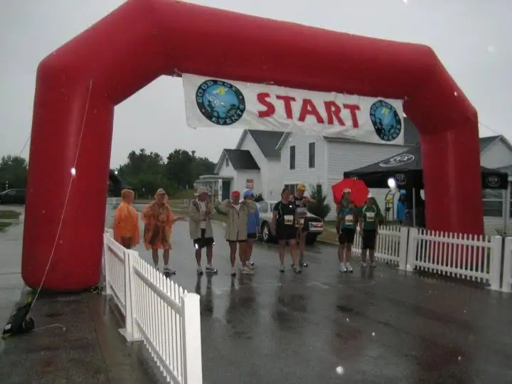 Rain-soaked runners gather beneath a red inflatable START arch beside a white picket fence.
