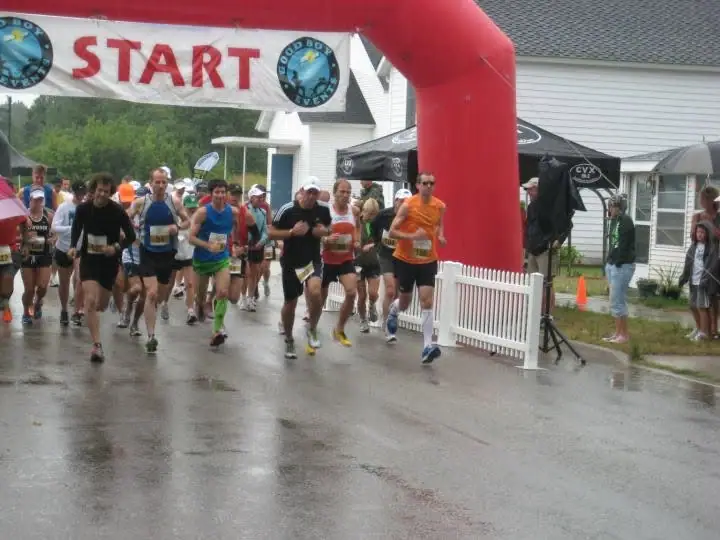 Runners begin a road race beneath a red START arch on a wet street as spectators watch.