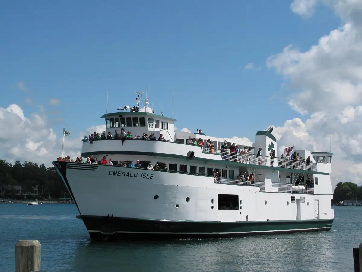 A crowded white multi-deck ferry travels past a wooded shore under a bright blue sky.