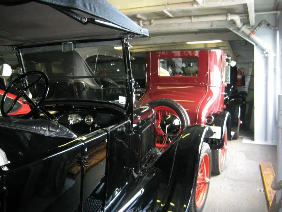 Two vintage automobiles, one black and one red, are parked indoors beneath exposed pipes and fluorescent lights.