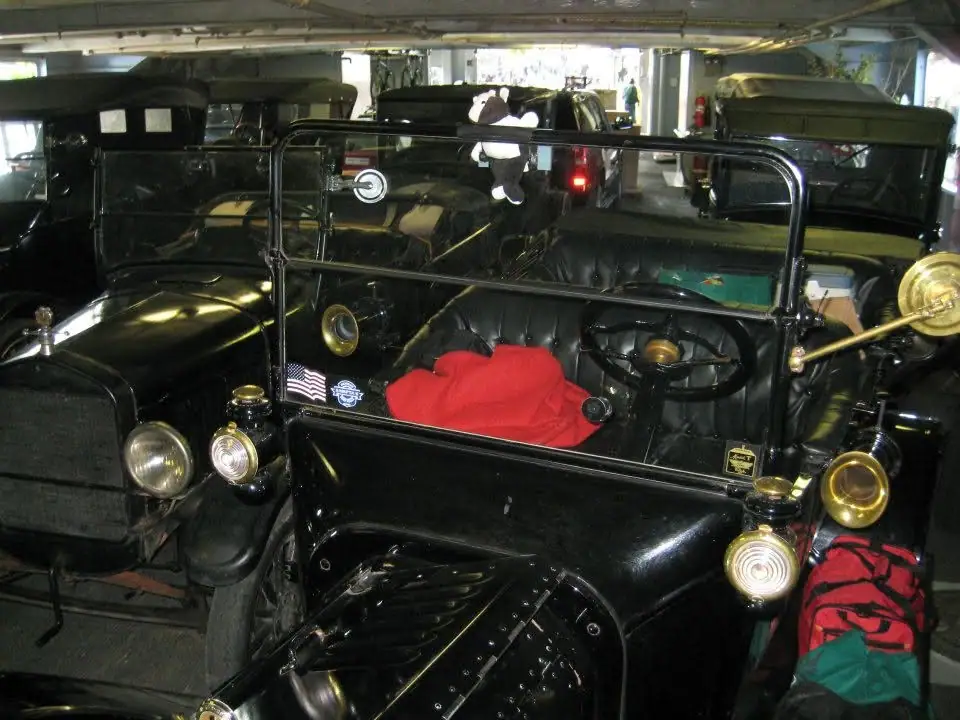 Black antique automobile displayed indoors among other vintage vehicles, with a red cloth on its seat.