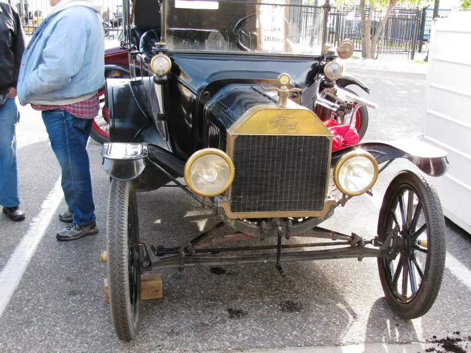 Front view of a black antique automobile with brass fittings, displayed outdoors beside two partially visible people.