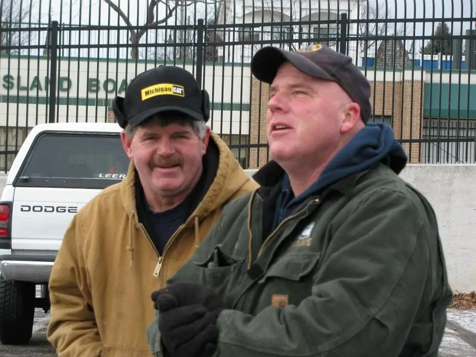 Two men in winter clothing stand outdoors beside a white pickup truck and a fenced building.