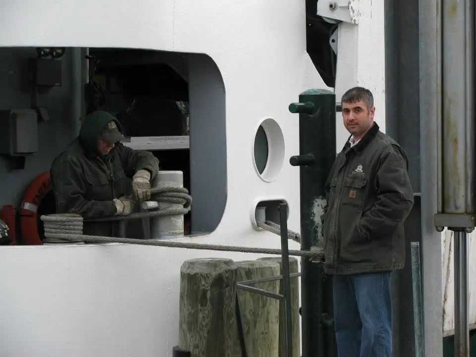 Two workers handle a thick mooring rope beside a ferry's white hull and deck equipment.