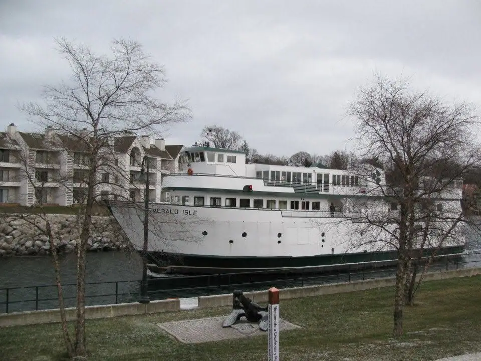 A white ferry named EMERALD ISLE moored beside a waterfront promenade with buildings and leafless trees.