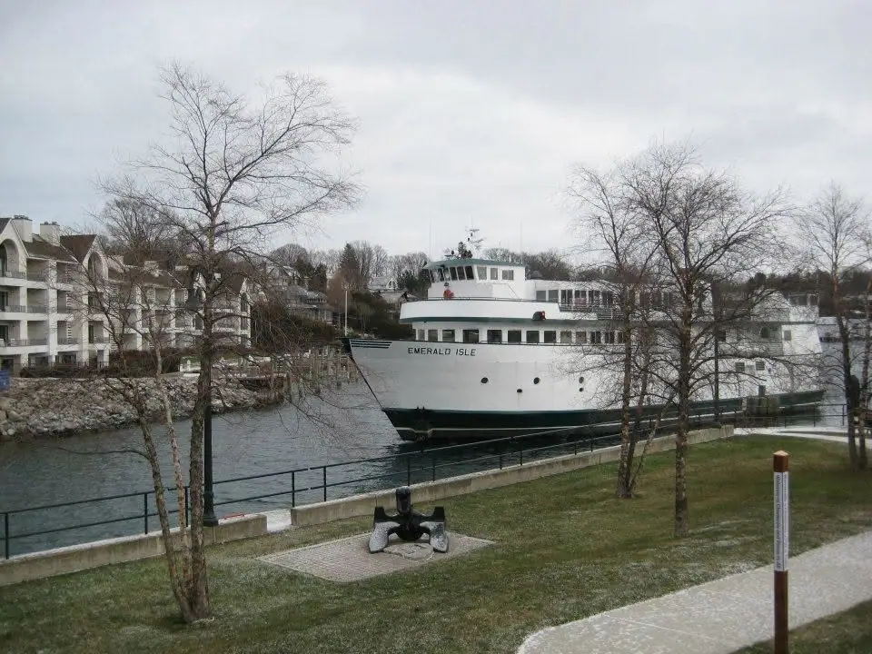 A white ferry moored beside a waterfront promenade, with an anchor sculpture and leafless trees in the foreground.