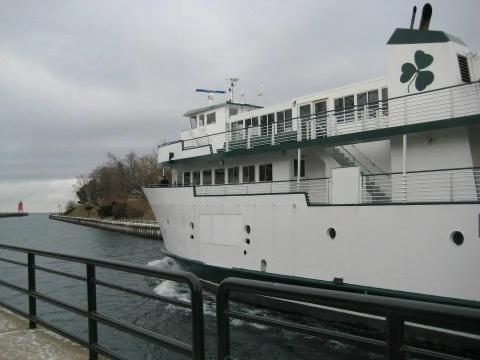 A white passenger ferry with a green shamrock emblem passes a waterfront railing beneath an overcast sky.