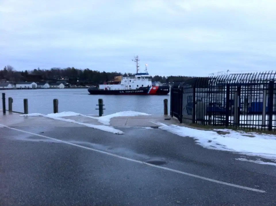 A Coast Guard vessel sits offshore beside a snowy dock, viewed through a tall metal fence.
