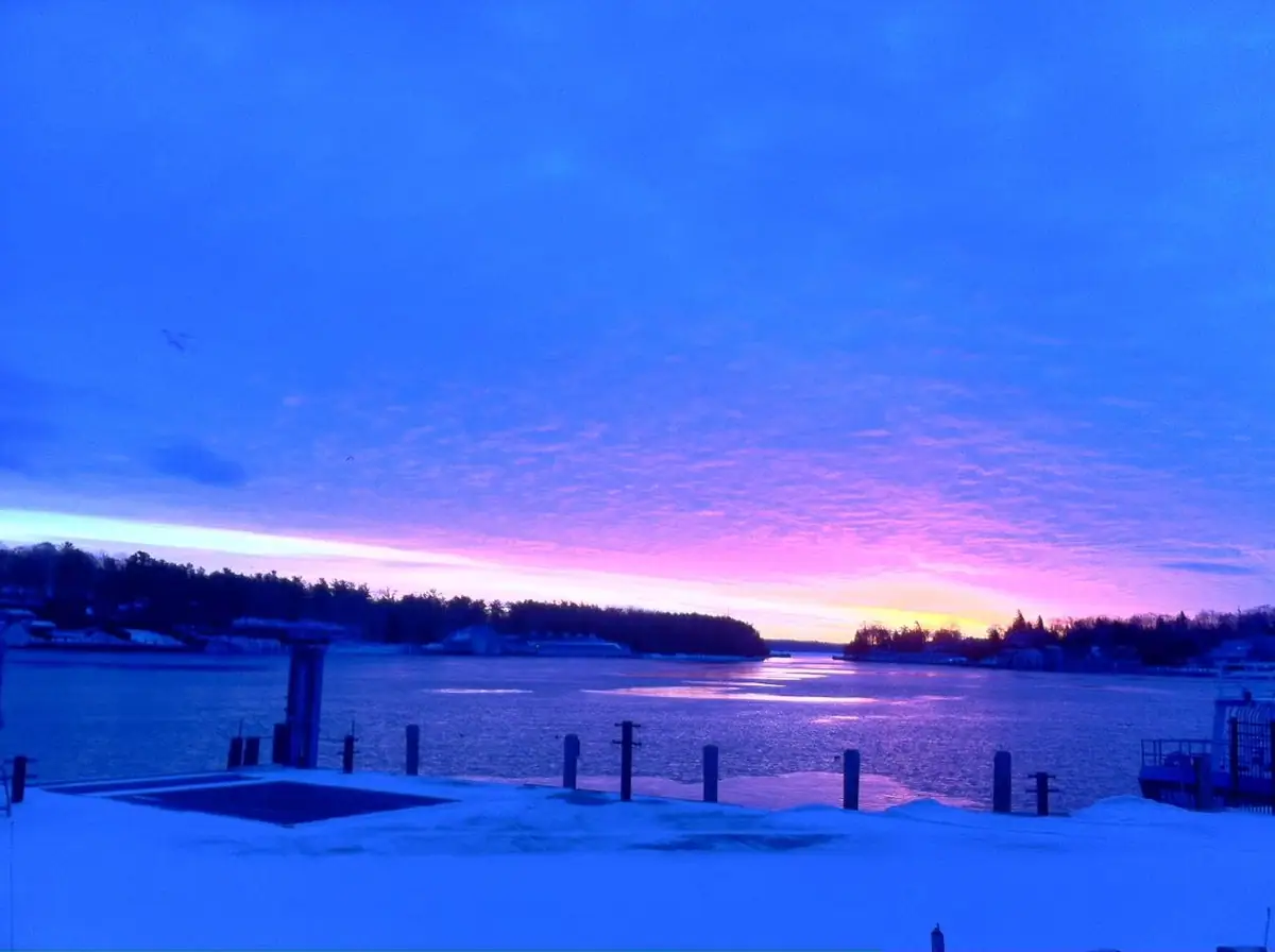 Sunrise or sunset colors the sky above a snowy waterfront with ice, pilings, and a tree-lined shore.
