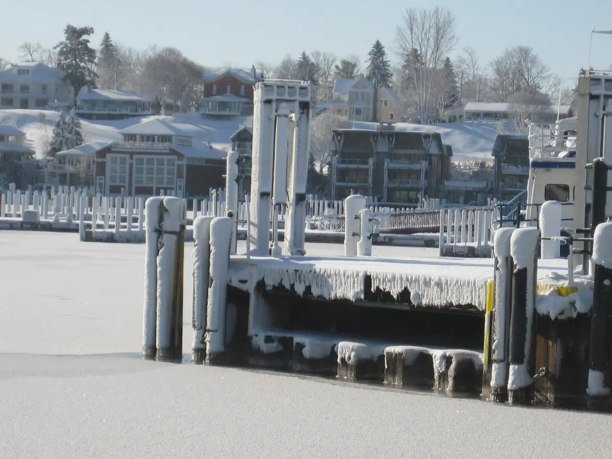 A snow-covered dock and ice-coated pilings extend across a frozen harbor, with houses and leafless trees behind.