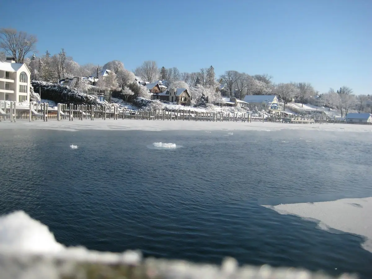 Snow-covered waterfront with rows of dock pilings, floating ice, houses, and trees beneath a clear blue sky.